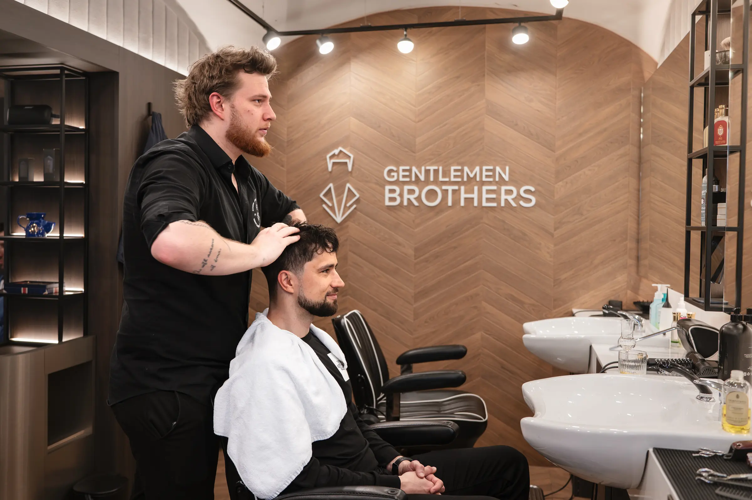 Holič Gentlemen Brothers upravuje zákazníkovi vlasy po střihu, v pozadí dřevěná stěna s logem